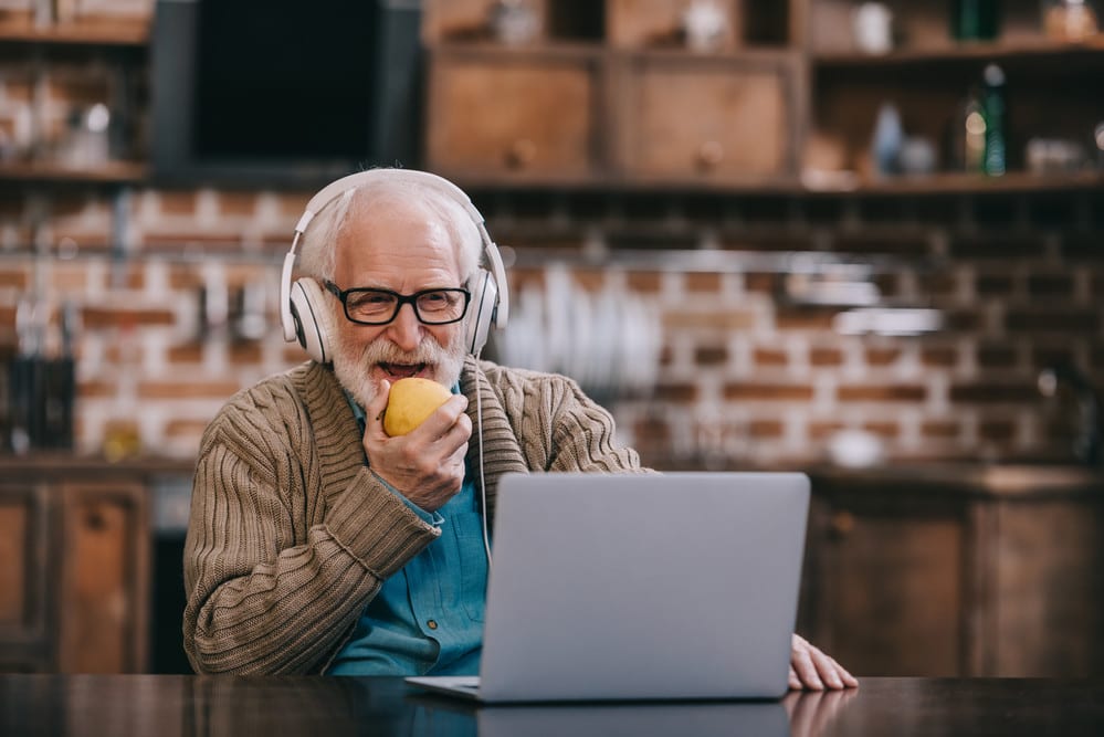 Senior man on laptop, smiling, eating apple