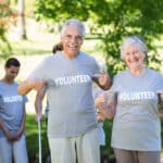 Two smiling seniors pointing at their t-shirts that read volunteer while standing in a park